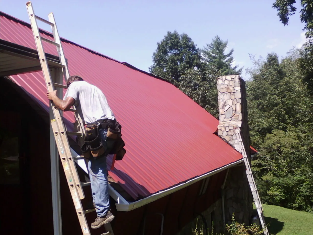 Roofer on a ladder beside a red metal roof and stone chimney — Soffit & Fascia Repair by Roofing Brothers in Ford City